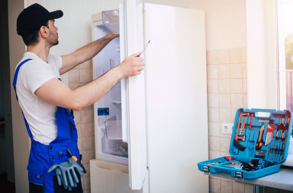 conserto-de-geladeira-rio-de-janeiro-profissional-jovem-reparador-com-uniforme-de-trabalhador-e-bone-com-caixa-de-ferramentas-moderna-com-equipamento-esta-consertando-a-geladeira-na-cozinha